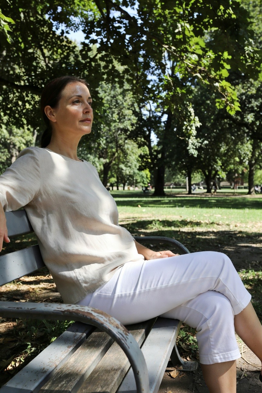 Woman in her 40s or 50s with brown ponytail wearing a loose cream short-sleeve blouse and white cropped straight-leg pants, seated sideways on a wooden park bench with one arm resting on the back, neutral expression looking off to the side