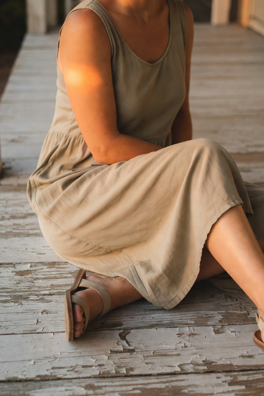 Woman sitting on wooden porch in sleeveless beige tank dress with gathered skirt and tan strappy flat sandals, legs extended casually