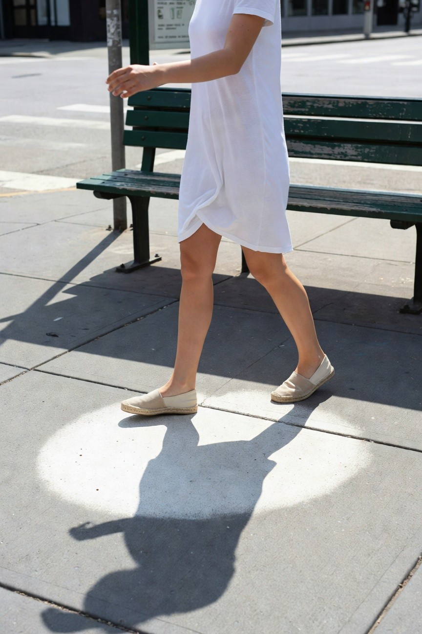 Woman walking in a short white short-sleeve dress and tan espadrille flats, casting a shadow on urban pavement near a green bench