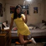 Woman in a bright yellow short-sleeved mini dress with waist tie, posing in front of a mirror while leaning on a wooden dresser, wearing white Converse sneakers, casual bedroom setting.