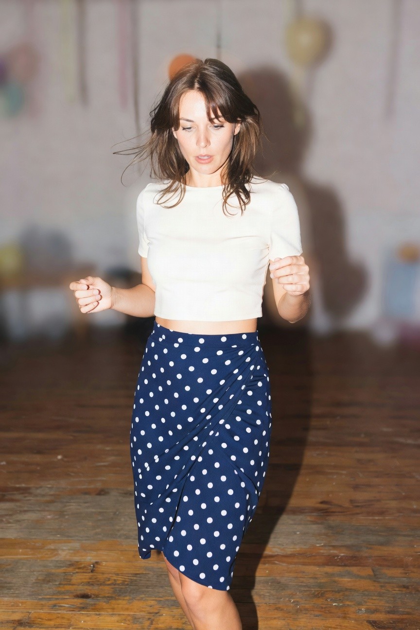 Woman in motion wearing a white cropped short-sleeve top and high-waisted navy blue skirt with large white polka dots, midriff exposed, in a studio setting