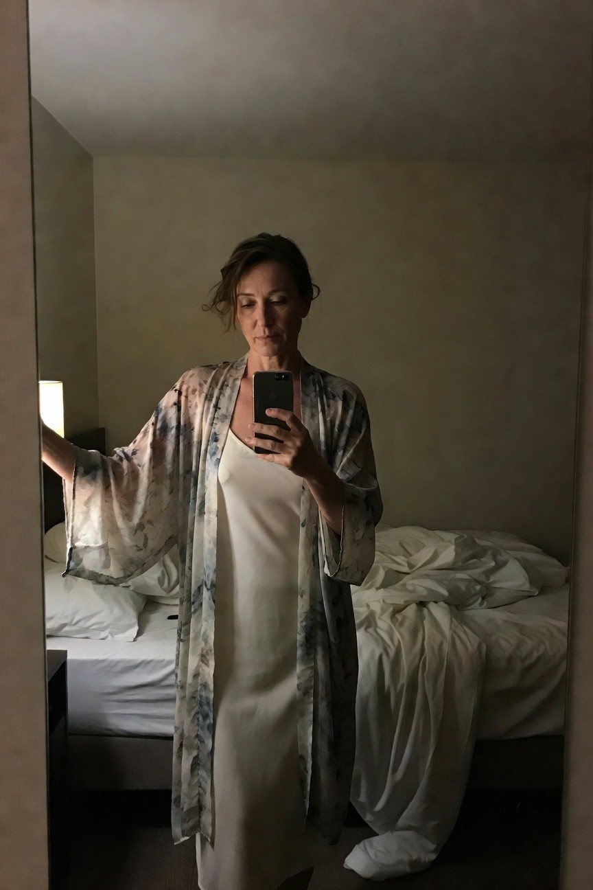 Mirror selfie of a woman in a hotel room wearing an open sheer floral kimono robe in blues and pinks over a white silk slip dress, wide sleeves extended, bed with white sheets in background