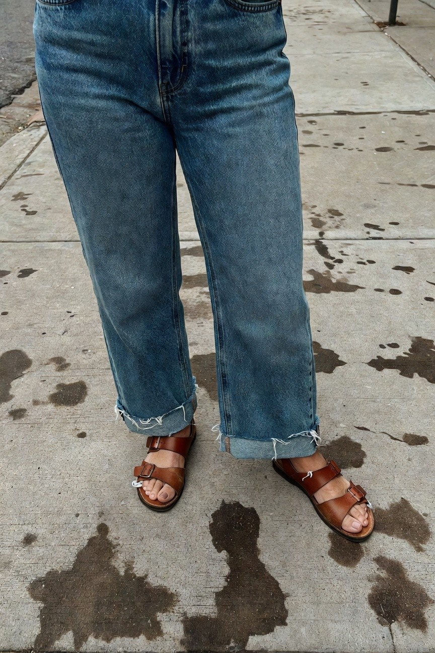 Close-up view of high-waisted light blue baggy jeans with frayed cropped hems paired with tan leather strappy sandals featuring buckles, standing on pavement