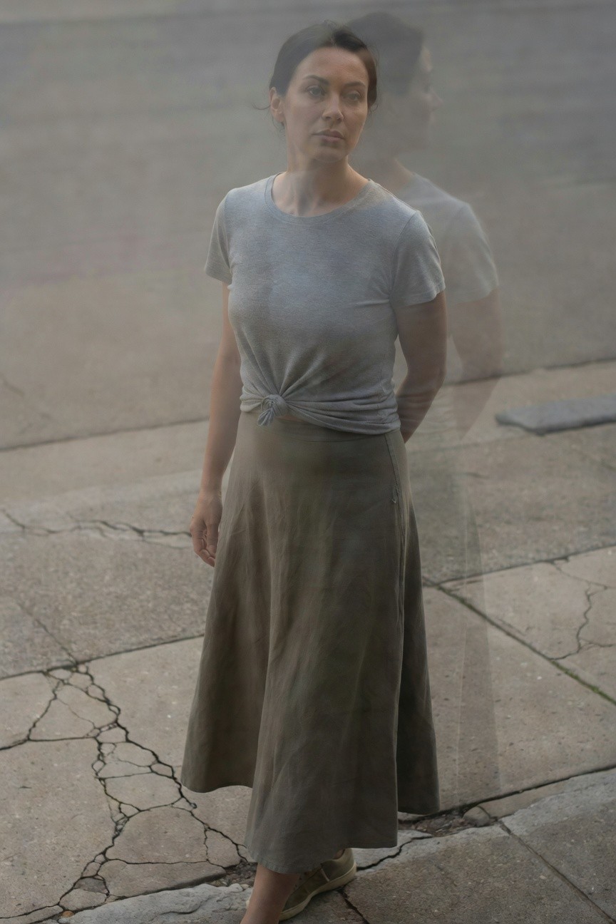 Woman in gray short-sleeve t-shirt knotted at waist paired with flowing khaki midi skirt and simple nude sandals, standing sideways on cracked pavement with foggy reflection