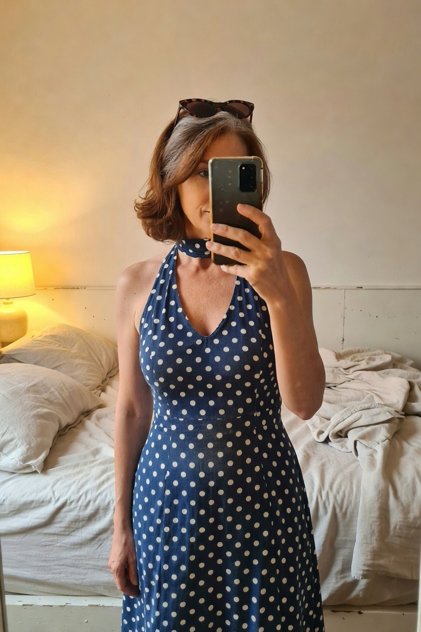 Mature woman with gray bob haircut wearing a fitted navy blue polka dot halter neck dress, sunglasses perched on head, taking a mirror selfie in a bedroom setting