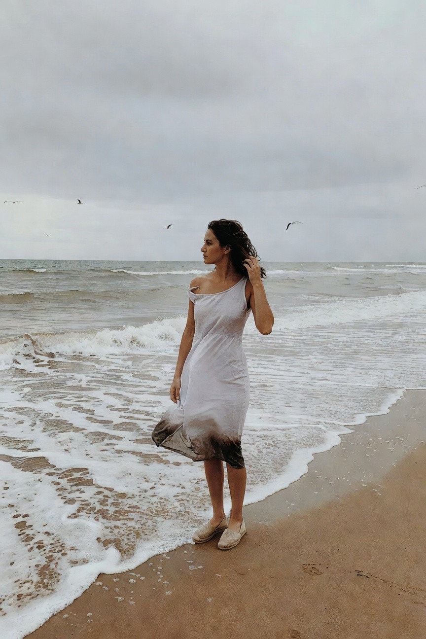 Woman standing on sandy beach in a flowy white one-shoulder midi dress with darker hem and tan espadrille wedge sandals, curly hair tousled, hand touching hair, ocean waves and seagulls in background
