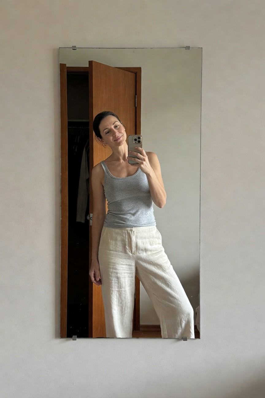 Woman taking a mirror selfie in a light gray sleeveless tank top and loose white linen pants, standing in front of an open wooden closet door on a plain wall.