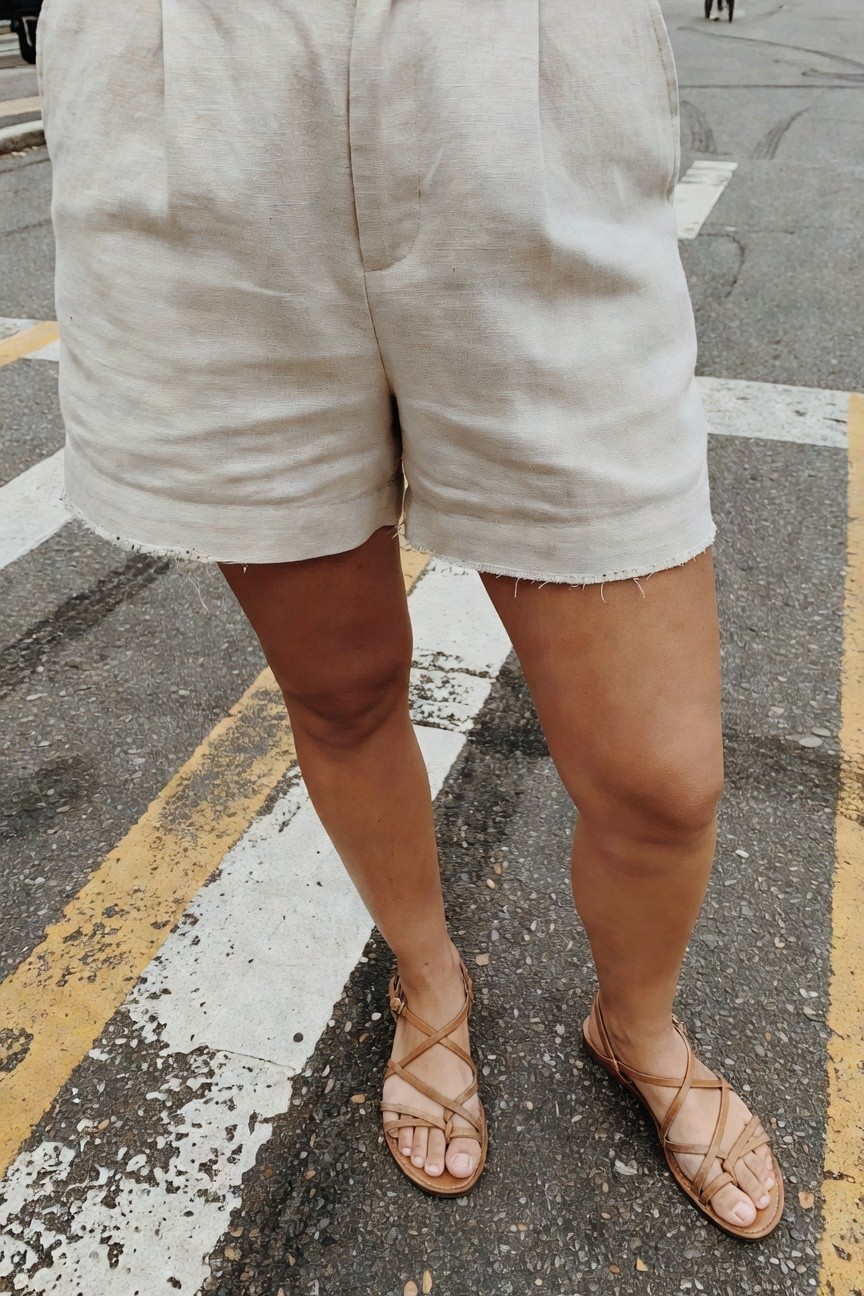 Close-up view of a woman standing at a crosswalk wearing light beige wide-leg linen shorts with pleated front and frayed hems paired with tan crisscross strappy flat sandals, focusing on legs and lower outfit against asphalt street.