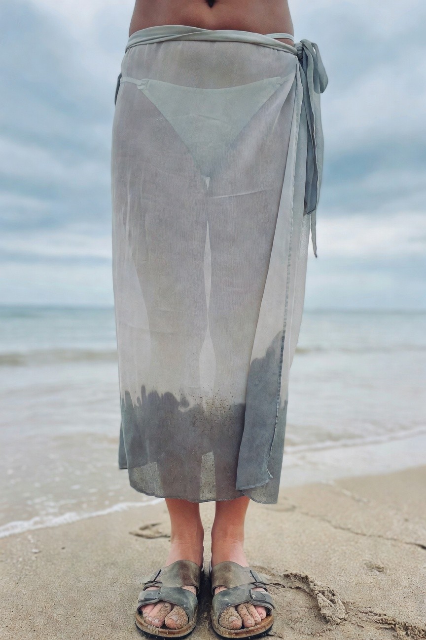 Woman standing in sheer pale gray wrap skirt tied at waist over matching pale bikini bottoms, wearing sandy tan Birkenstock sandals, sand visible on legs and feet