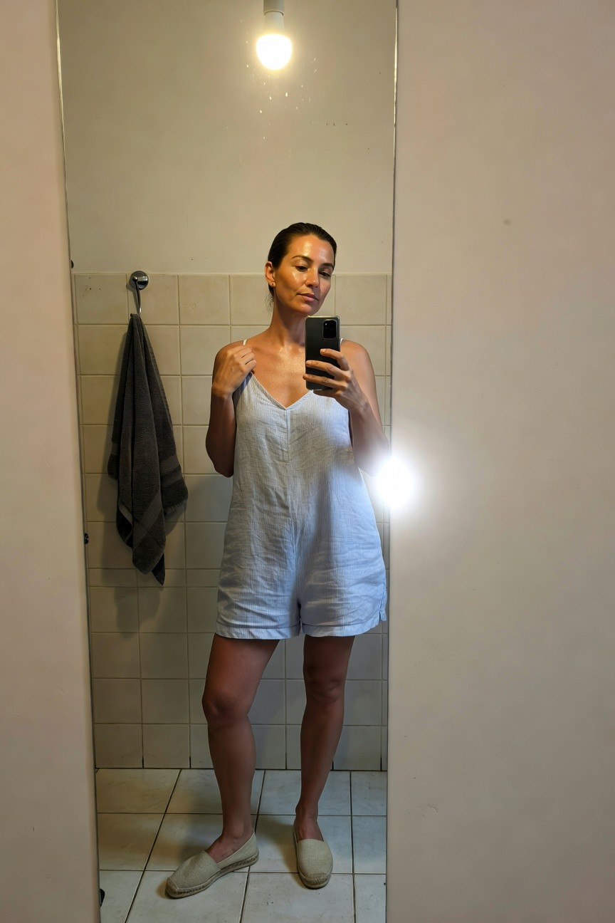 Woman taking a mirror selfie in a bathroom, wearing a light blue linen spaghetti-strap romper with short legs, nude espadrilles, hair in a bun, gray towel hanging nearby