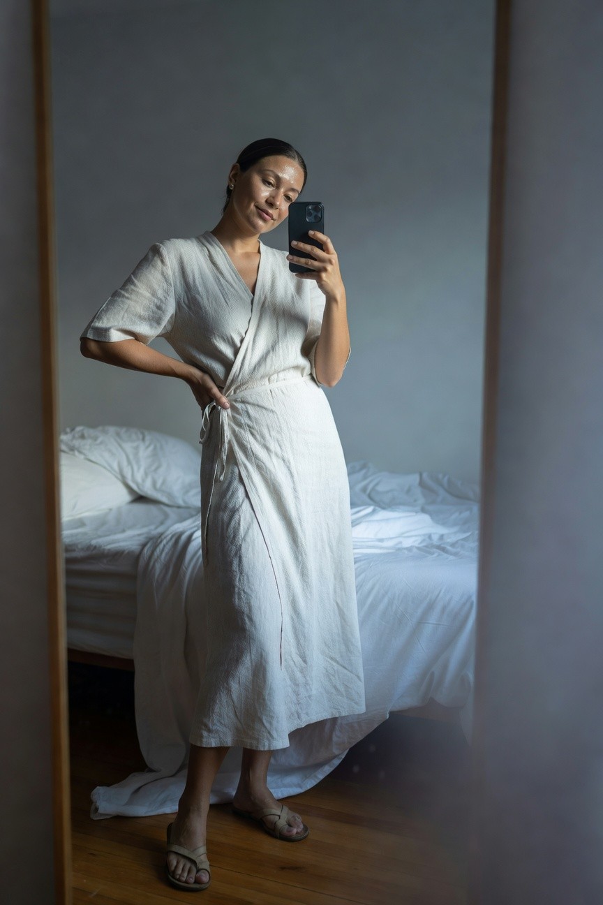 Woman taking mirror selfie in a white linen wrap dress with short sleeves and tie belt, paired with tan flat sandals, standing in a bedroom with bed visible