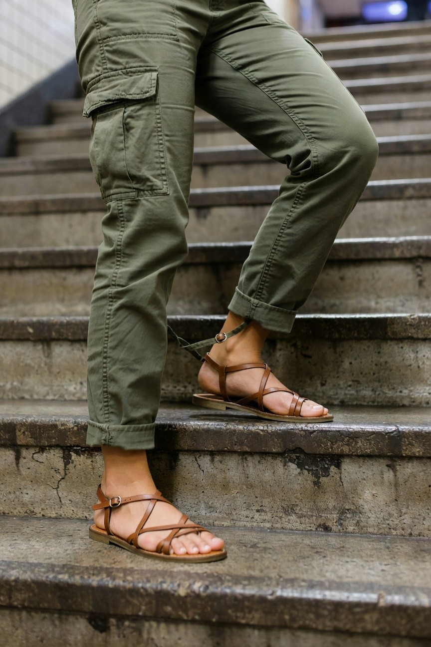 Close-up view of a woman's lower legs and feet on urban concrete stairs, wearing loose olive green cargo pants with rolled cuffs and multiple pockets, paired with tan leather crisscross strappy flat sandals with ankle straps