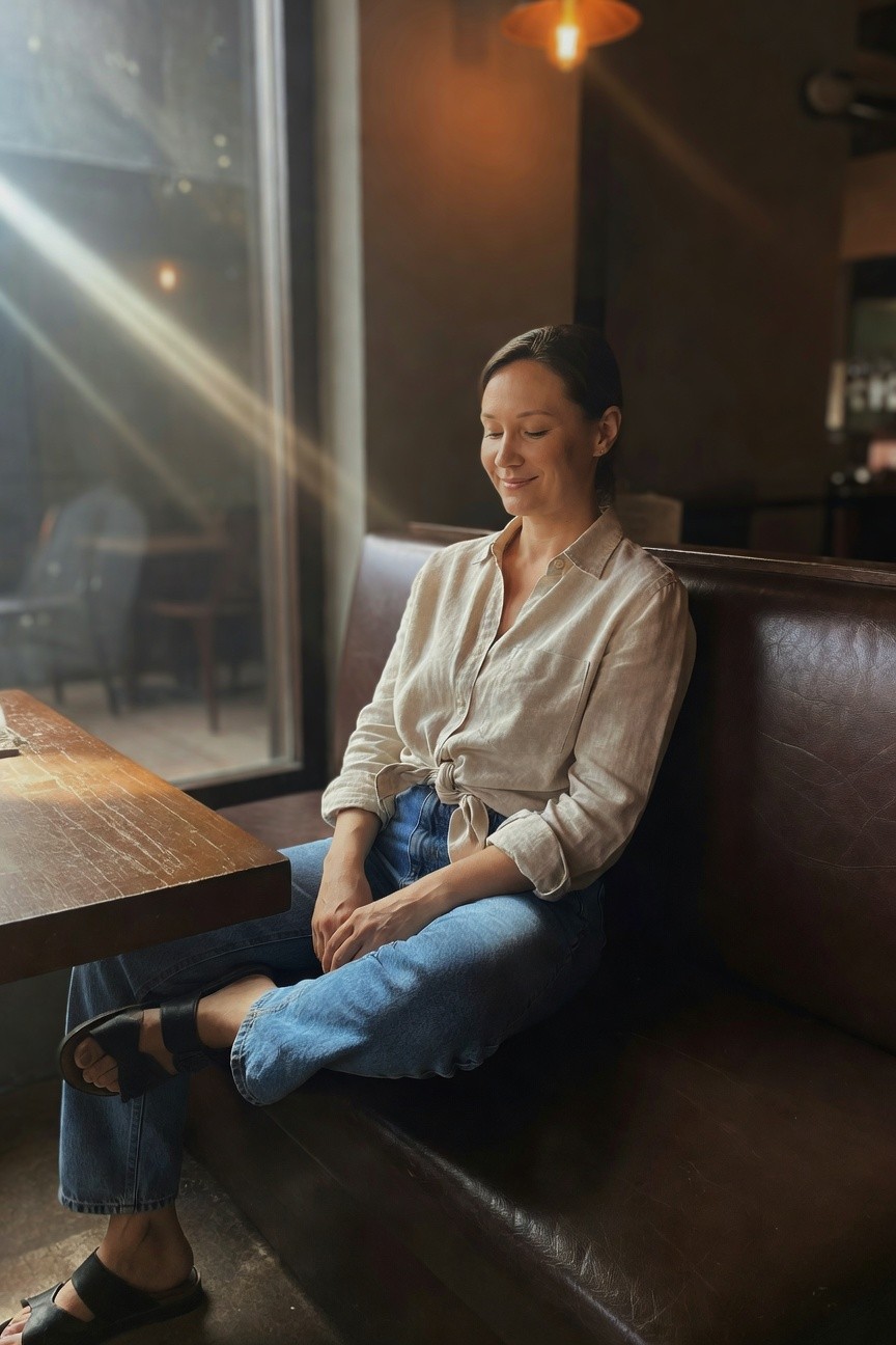 A woman in a knotted beige shirt and blue jeans sits in a dimly lit café.