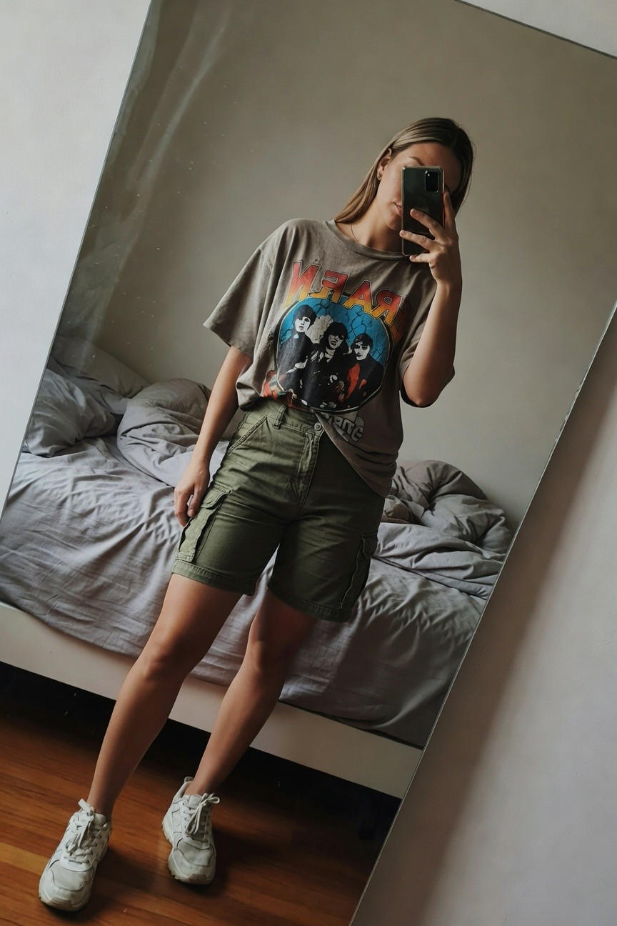 Mirror selfie of a woman in an oversized gray Metallica band t-shirt with colorful graphic, olive green mid-thigh cargo shorts cinched with a belt, and white chunky sneakers, posing in a simply furnished bedroom with bed visible in mirror