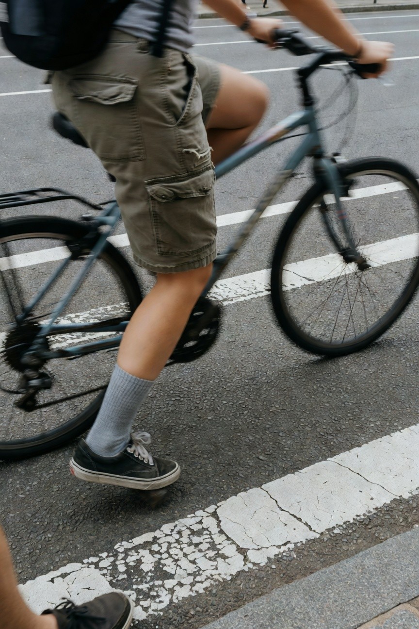 Close-up of a person biking in urban bike lane wearing loose khaki cargo shorts with pockets, gray tank top, black backpack, gray crew socks, and black lace-up Vans sneakers, motion blur on wheels
