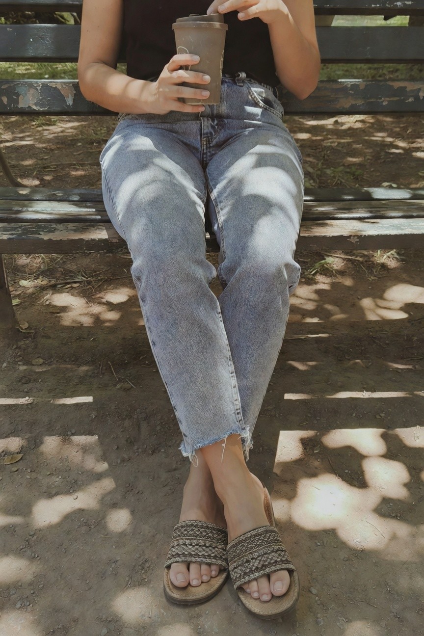 Woman sitting on a bench holding a brown to-go coffee cup, wearing a black cropped tank top, light wash distressed baggy jeans cropped at the ankle, and tan braided slide sandals, casual summer outfit