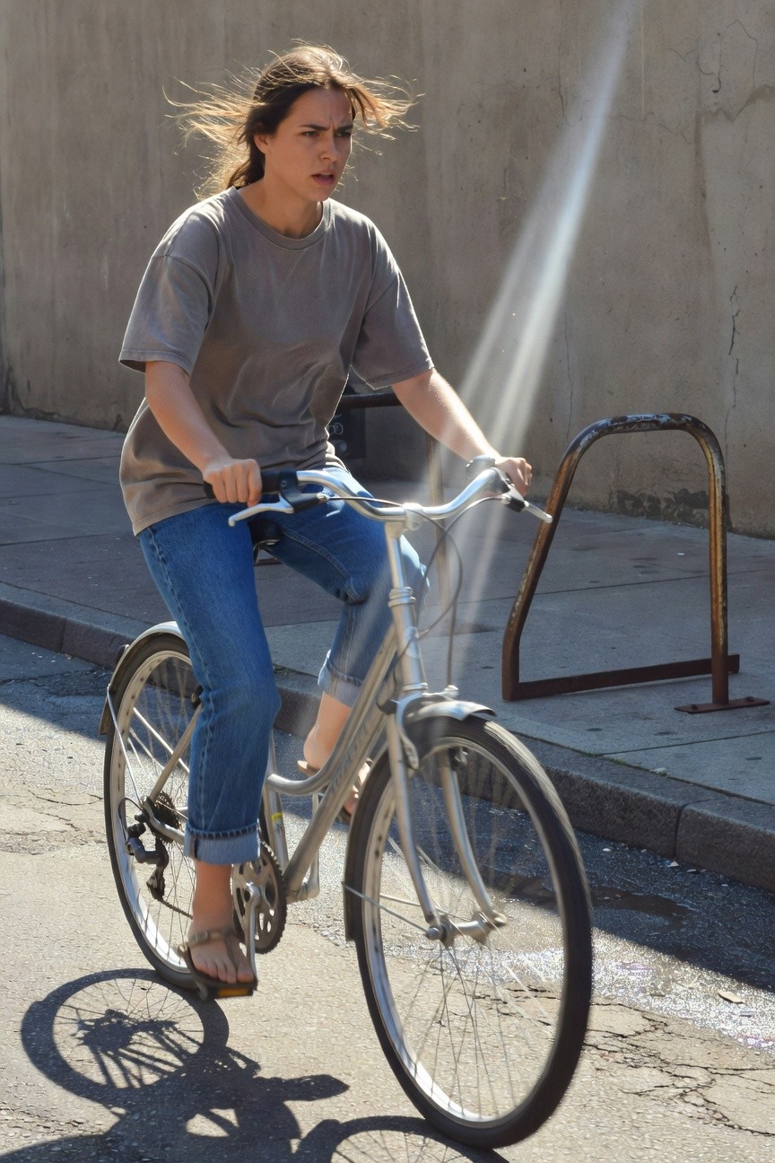Woman biking in oversized gray short-sleeve t-shirt, light blue straight-leg jeans cuffed at ankles, tan flip-flop sandals, silver city bike on sunny street.