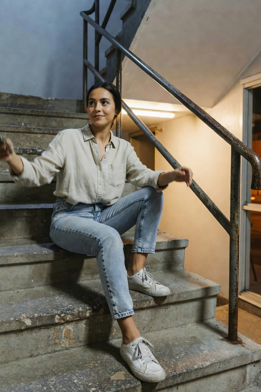 Woman seated on concrete stairs in a loose beige long-sleeve linen button-down shirt with rolled cuffs, light wash blue straight-leg jeans, and white low-top sneakers, arms gesturing casually.