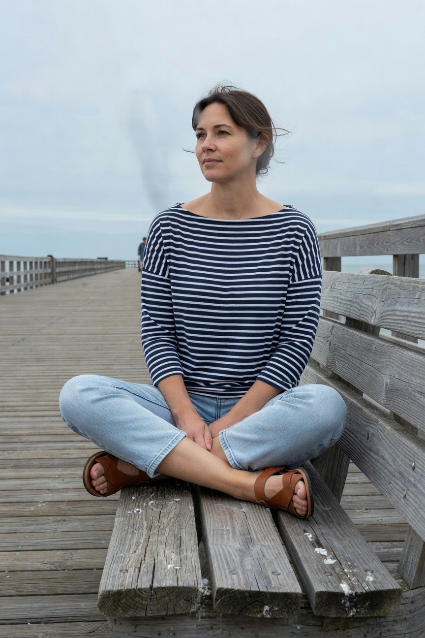 Woman in her 40s with brown hair in a loose bun sits cross-legged on a wooden bench wearing a navy and white horizontal striped boatneck long-sleeve top, light blue jeans, and tan leather strap sandals, looking thoughtfully to the side on a pier