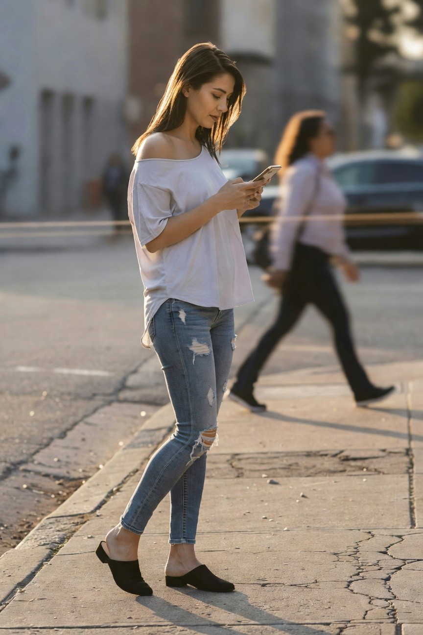 Woman in white off-shoulder top, light blue slim-fit ripped jeans with distressing at thighs and knees, and black low-heel mule slides, standing casually while checking phone