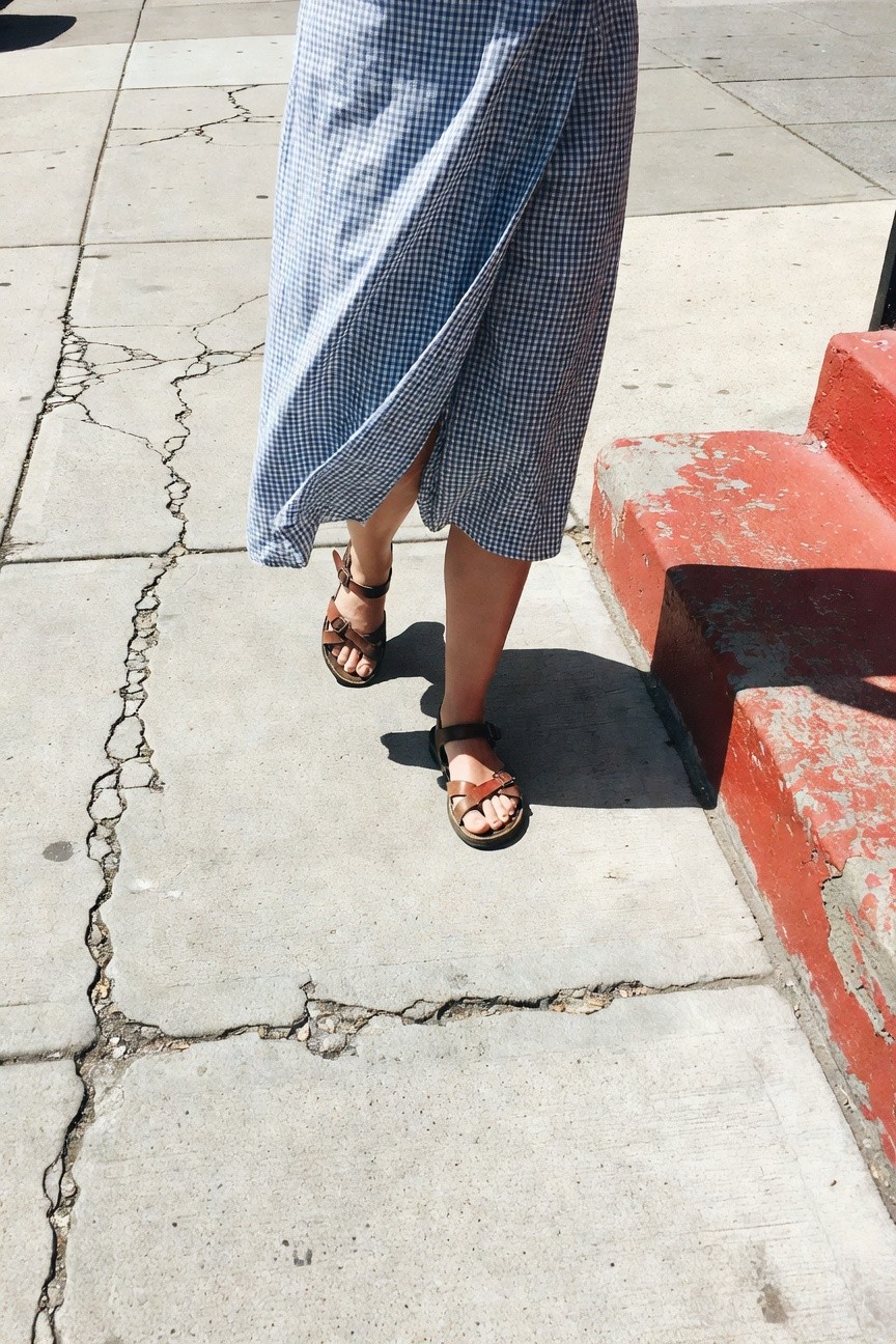 Woman walking on cracked sidewalk in a blue-and-white gingham wrap dress with brown leather strappy sandals, red steps nearby