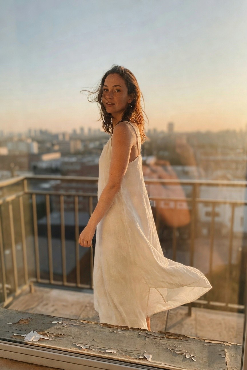 Woman standing on a rooftop balcony in a flowing off-white linen spaghetti-strap slip dress reaching mid-calf, turning slightly with wind lifting the hem, cityscape in background at dusk