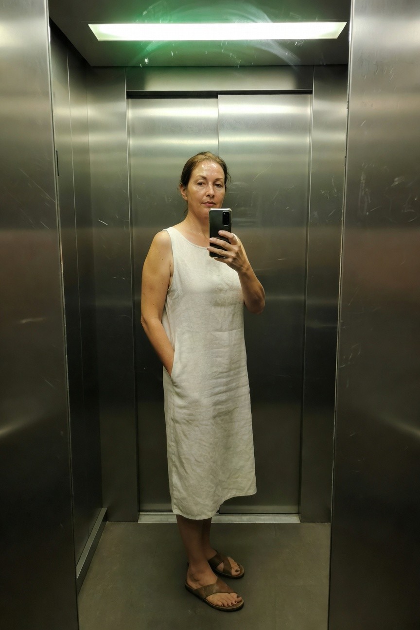 Woman in her 50s taking a selfie in an elevator mirror, wearing a sleeveless white linen midi sundress with hands in pockets and brown strappy flat sandals