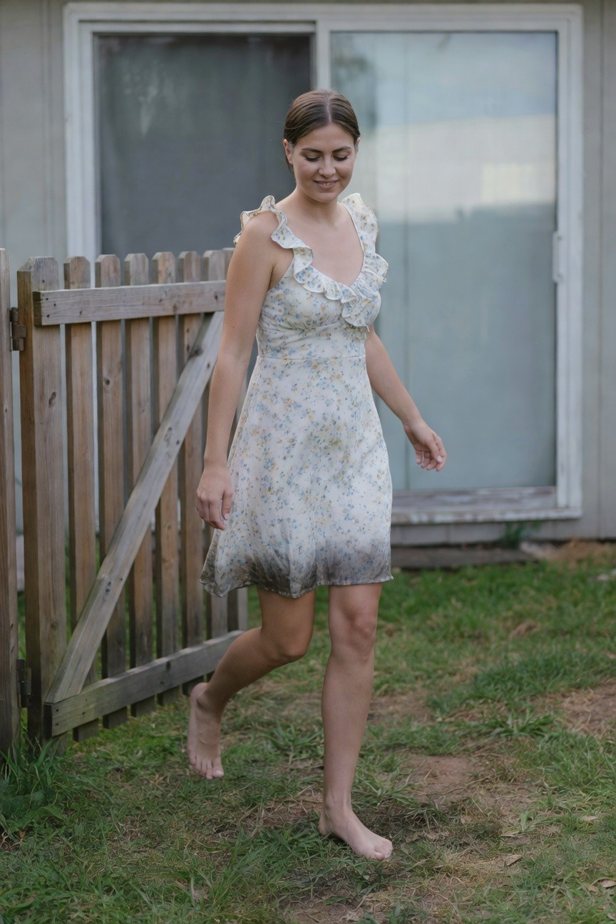 Woman walking barefoot in a backyard, wearing a short ruffled sundress with small floral print in light colors, fading darker at the hem, against a wooden fence and house siding.