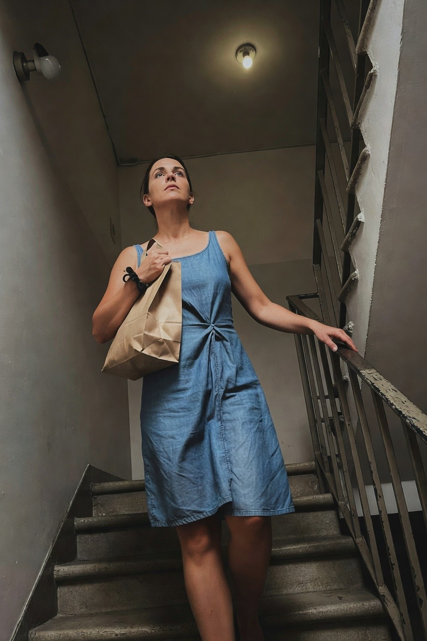 Woman in a sleeveless blue denim sundress with knotted waist, holding a brown paper bag, ascending concrete stairs in a dimly lit indoor stairwell, hand on metal railing
