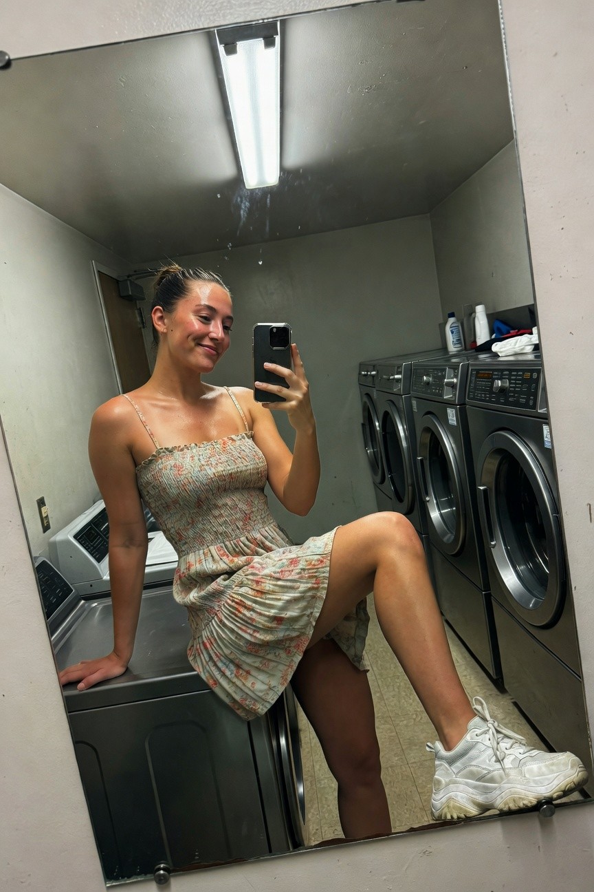 Woman smiling in selfie wearing short smocked floral sundress with spaghetti straps and white chunky sneakers, sitting on washing machine in laundry room