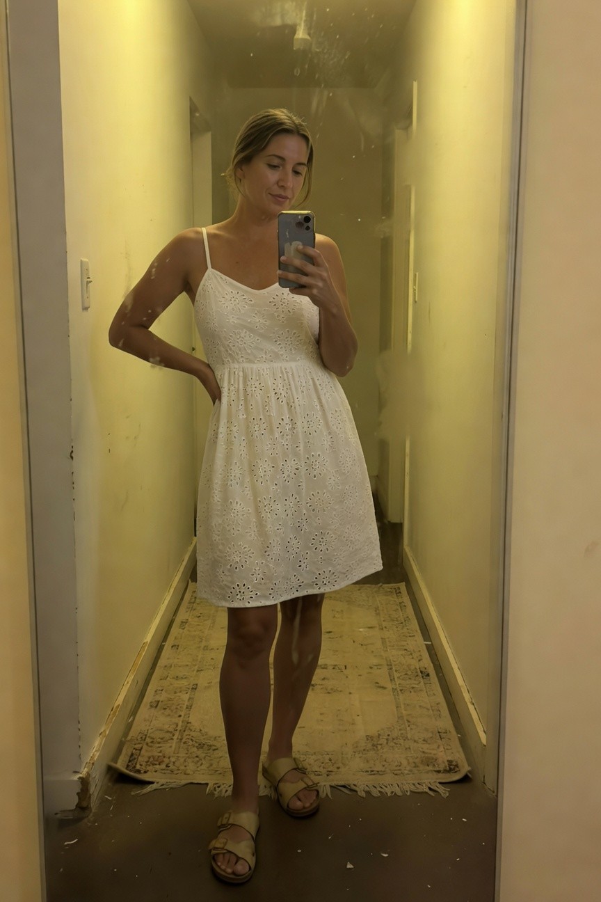 Woman taking a mirror selfie in a hallway wearing a white spaghetti-strap eyelet sundress with a fitted bodice, flared mid-thigh skirt, and tan buckled strappy sandals, one hand on hip.