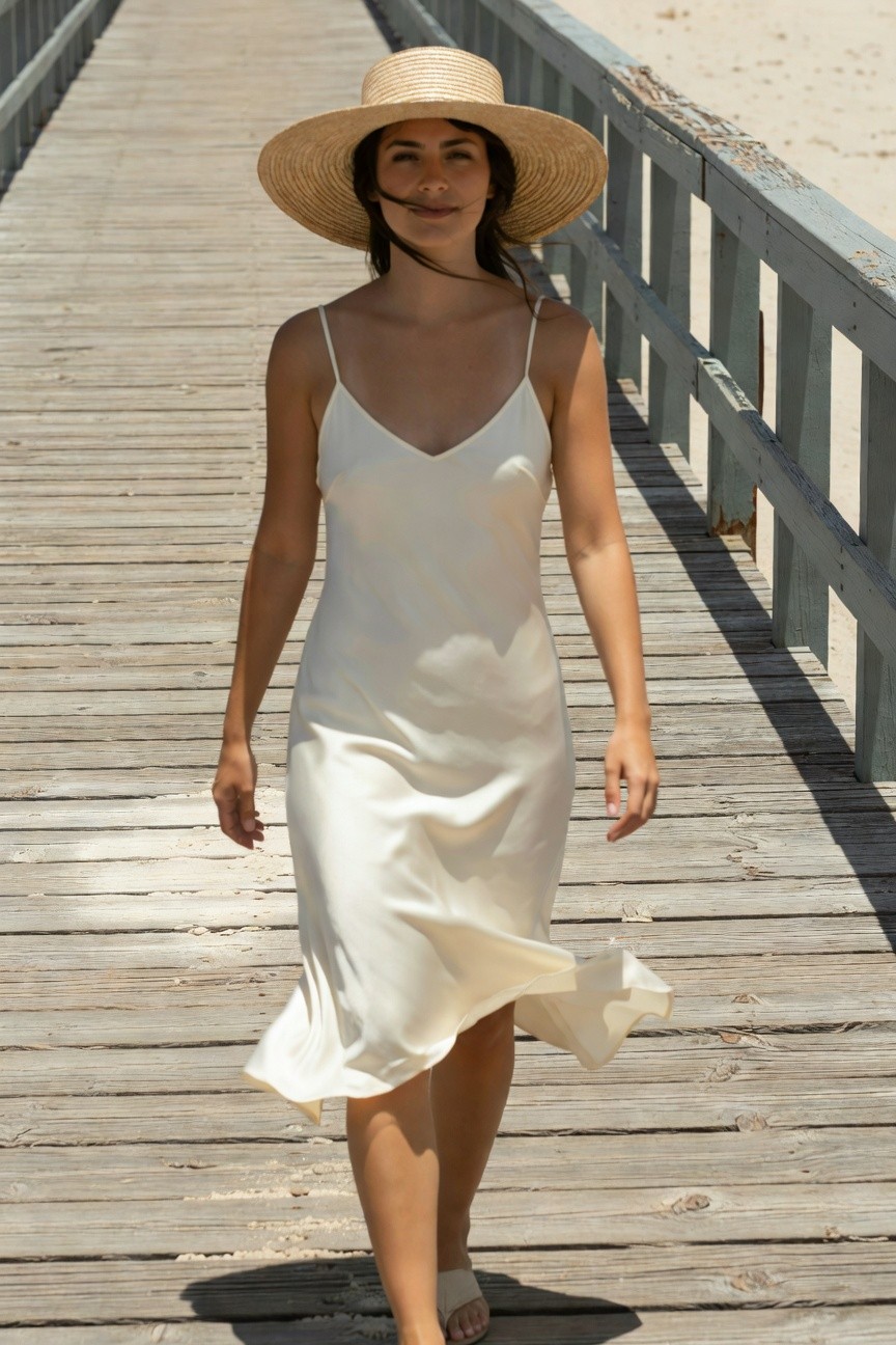 Woman walking on wooden pier in white silk spaghetti-strap slip dress with flared hem, wide-brim straw hat, and white strappy flat sandals, dark hair flowing