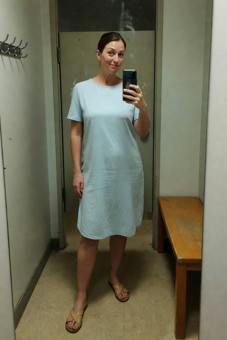 Woman in her 40s taking a mirror selfie in a changing room while wearing a loose-fitting light blue short-sleeve midi shift dress and tan cross-strap flat sandals, standing casually with one hand on hip