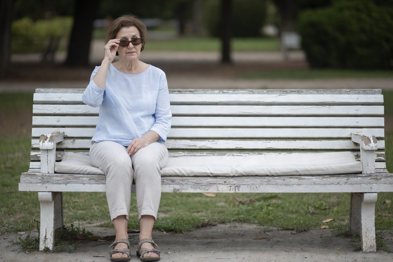 Mature woman seated on a white park bench wearing a light blue long-sleeve linen blouse, beige linen pants rolled at ankles, brown strappy sandals, and dark sunglasses lifted to her forehead, posed casually with hands in lap