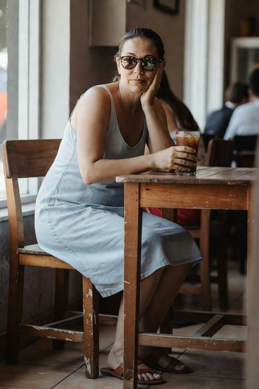 Woman with dark hair in tortoiseshell sunglasses and light blue sleeveless linen dress sitting at wooden cafe table holding iced coffee glass, tan flat sandals on feet, hand resting on cheek