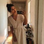 Woman with brunette hair in a messy bun taking a selfie in a pale beige short-sleeve linen wrap dress with waist tie, standing barefoot in a hallway next to a wooden shoe rack filled with sneakers