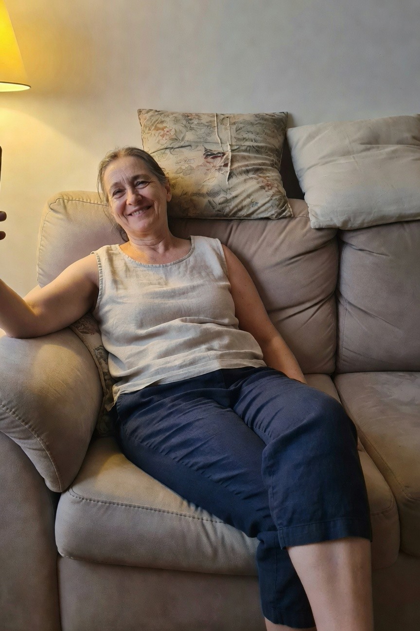 Woman in her 50s smiling selfie on beige sofa, wearing sleeveless beige linen tank top and navy blue cropped linen pants, arms relaxed, gray hair loose