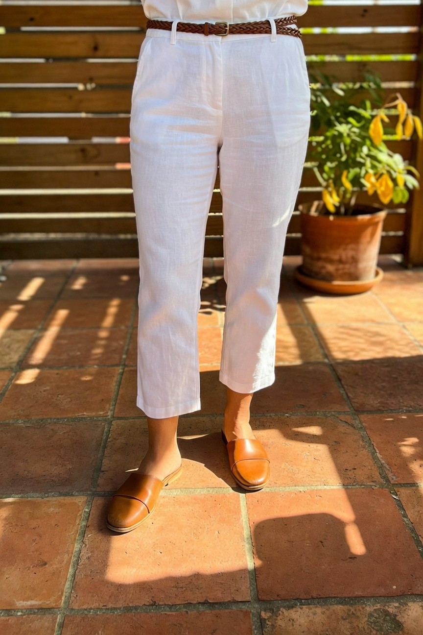 Woman modeling cropped wide-leg white linen pants belted with tan braided leather belt over tucked white linen blouse, paired with tan leather slide mules, standing on tiled patio