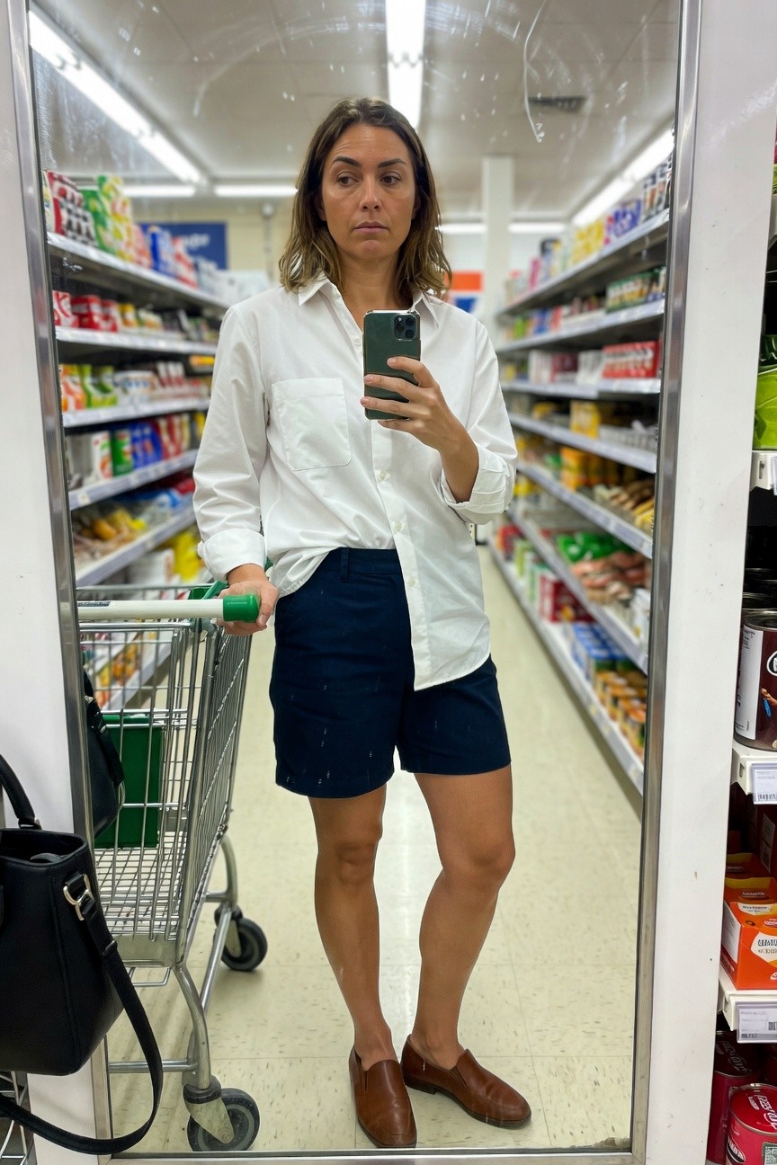 Woman taking mirror selfie in supermarket aisle wearing loose white button-up shirt, navy knee-length shorts, brown leather loafers, black handbag, and holding green shopping basket in metal cart