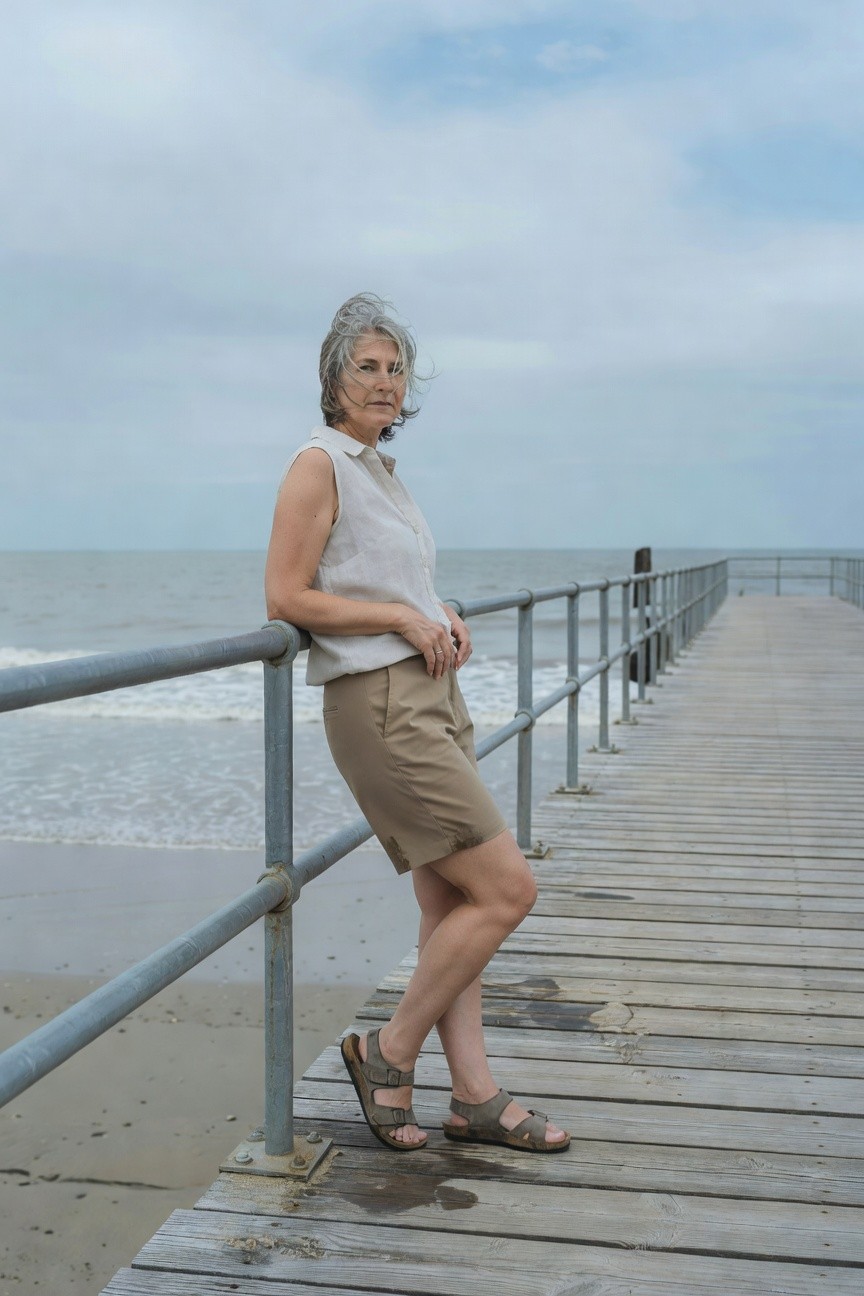 Silver-haired woman in her 50s or older wearing a white sleeveless collared blouse, khaki tailored shorts, and tan leather flat sandals, leaning casually on a metal pier railing with beach in background