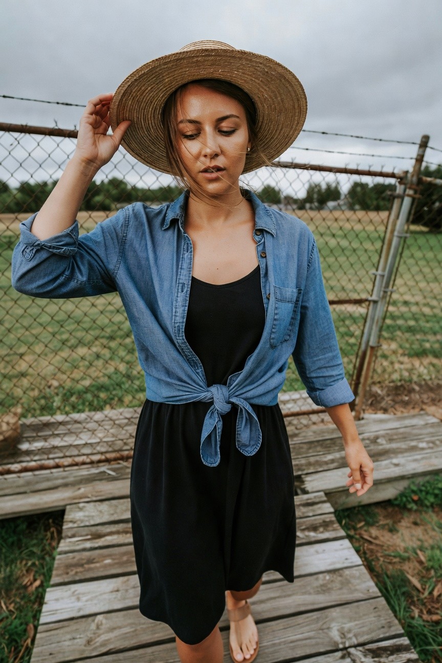 Woman in a wide-brim straw hat, open light blue denim shirt tied at the waist over a black spaghetti-strap midi dress, walking barefoot on a wooden path, hand adjusting hat