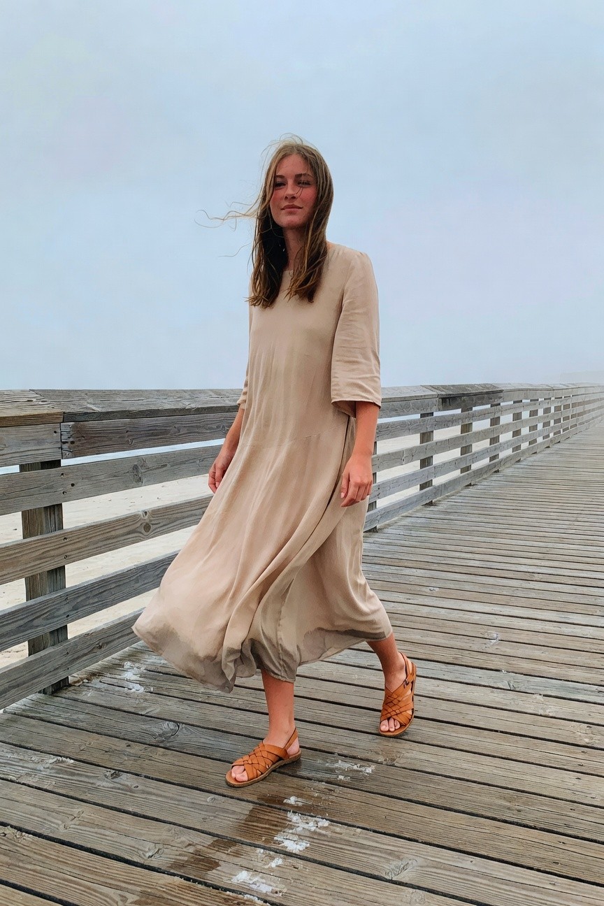 Woman walking on a wooden pier in a loose-fitting beige maxi dress with wide sleeves and flowy skirt, paired with tan braided strap sandals, hair windswept.