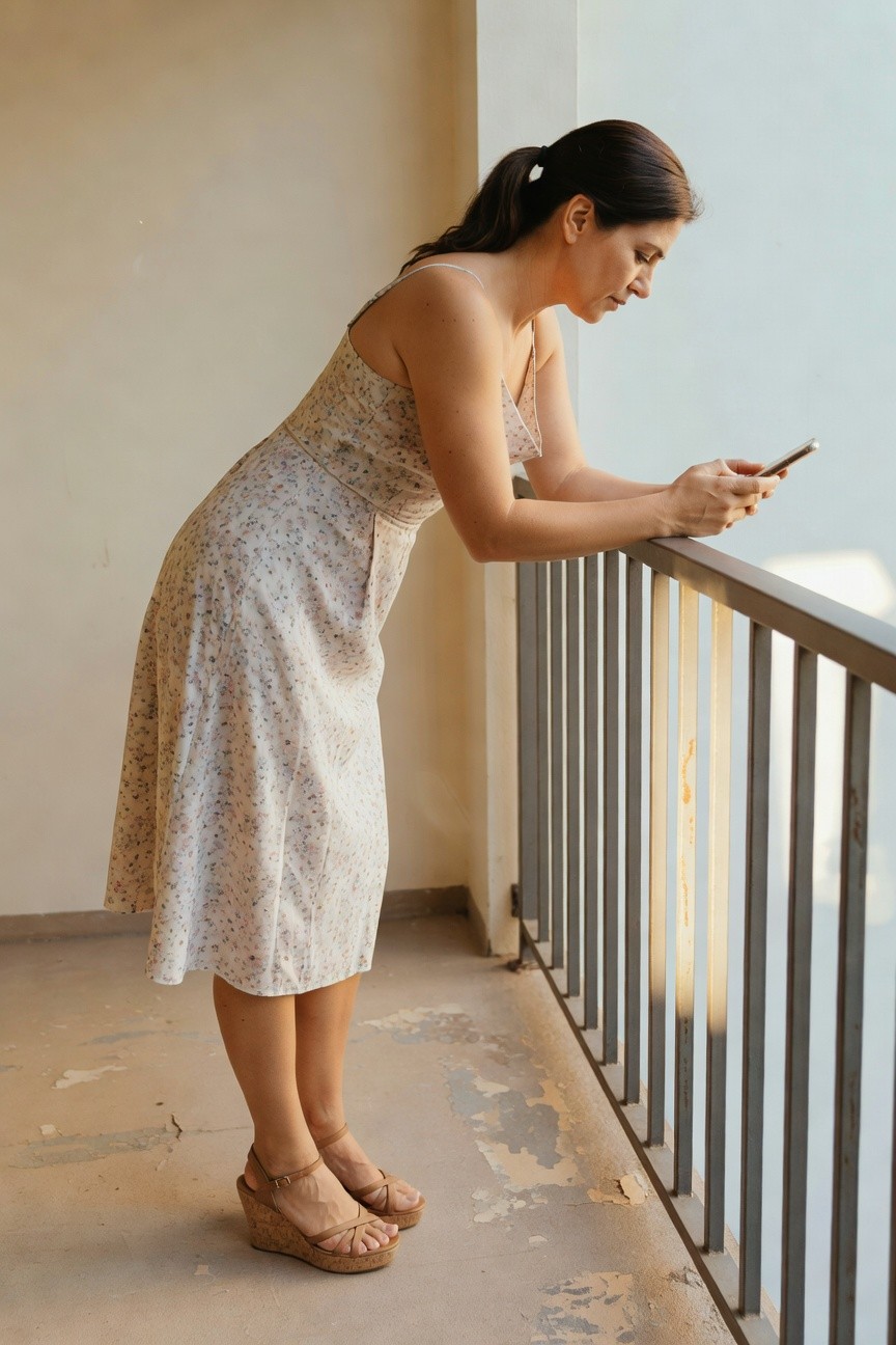Woman in a floral dress leaning on a balcony railing, looking at her phone.