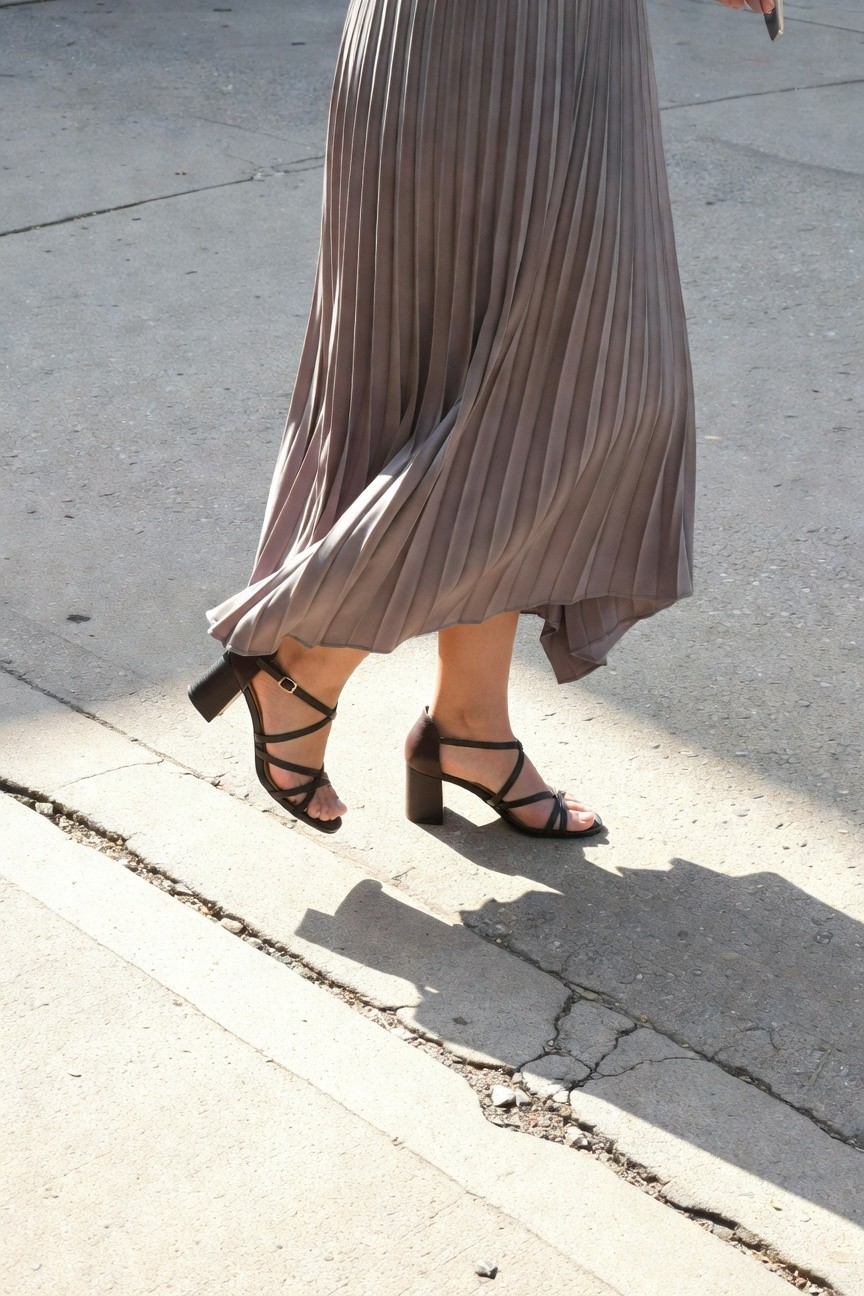 Woman walking in a silvery gray pleated midi skirt and black crisscross strappy block-heel sandals, holding a phone, on a concrete sidewalk