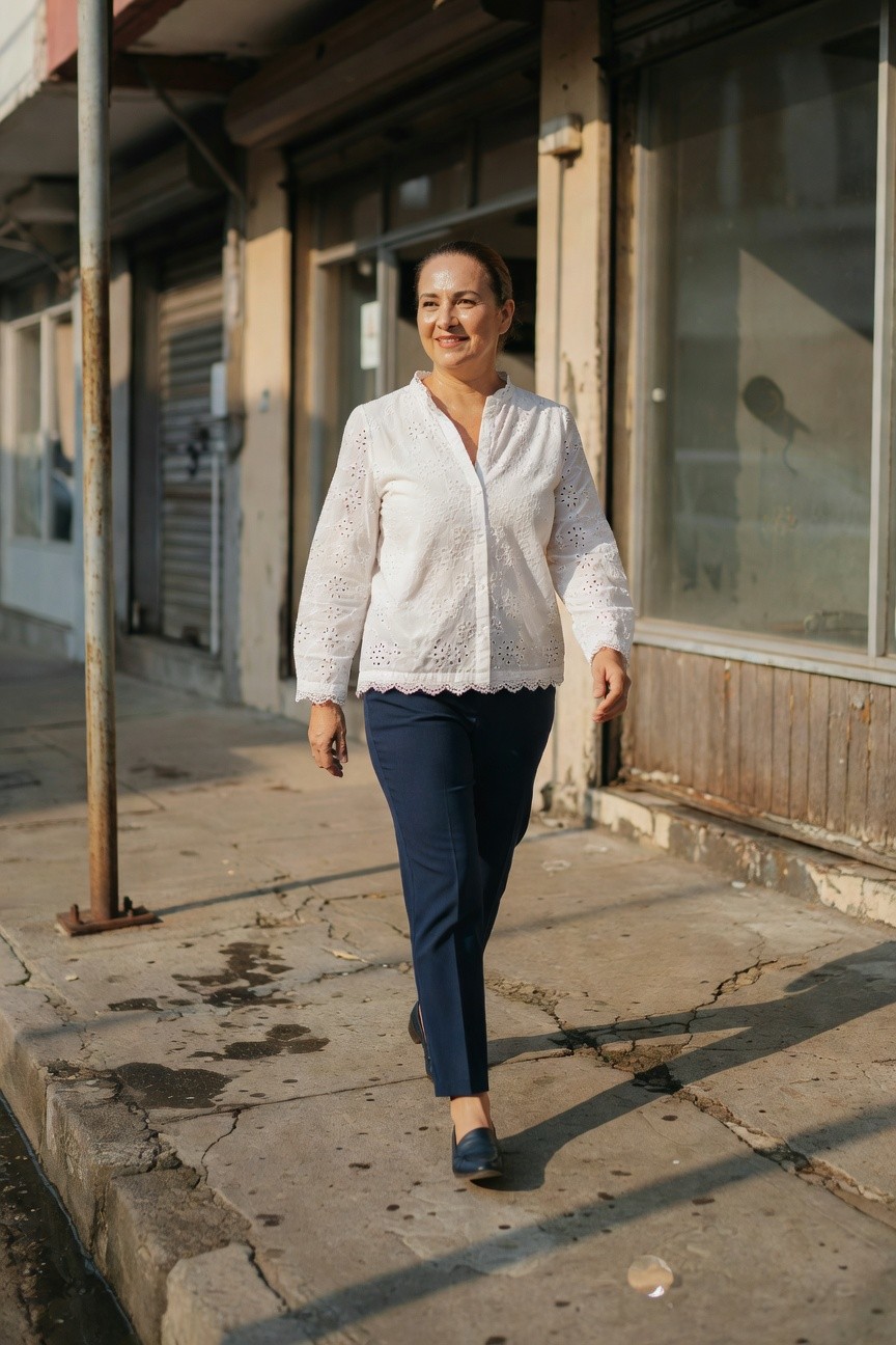 Middle-aged woman in a white eyelet-embroidered long-sleeve blouse with scalloped hem, slim navy trousers, and navy loafers, walking on a cracked sidewalk