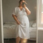 Plus-size woman in a white linen wrap dress with short sleeves and V-neck, holding the skirt fabric, wearing tan strappy sandals, taking a mirror selfie in a bedroom.