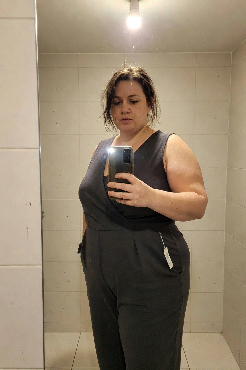Plus-size woman taking a mirror selfie in a dark gray sleeveless wrap jumpsuit with wide legs and pleated waist details, standing in a tiled bathroom