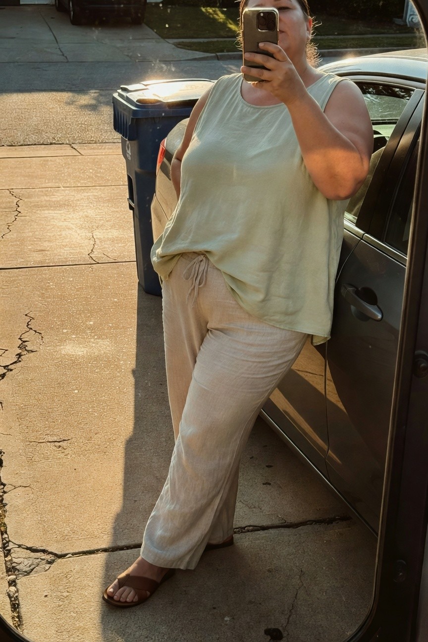 Plus-size woman in a loose light green sleeveless tank top and wide-leg beige linen drawstring pants with brown sandals, posing casually by a car door