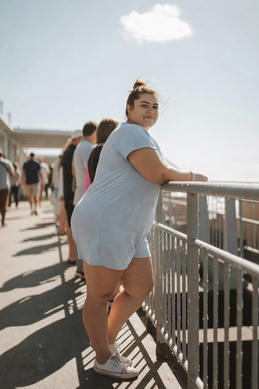 Plus-size woman wearing a short light blue short-sleeved dress leaning against a railing with white sneakers and a bracelet