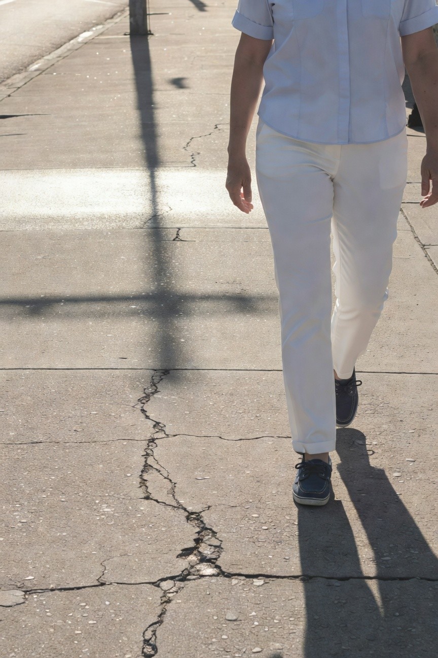 Woman in light blue short-sleeve chambray button-up shirt tucked into straight-leg white trousers, finished with navy boat shoes, walking on a cracked sidewalk with long shadows